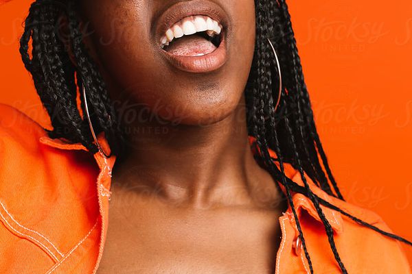 Woman wearing orange clothing standing behind orange wall holds hand over eyes and opens mouth 