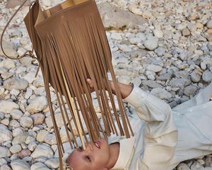 Model laying on a rocky beach holding up a tan fringed REE Projects handbag.
